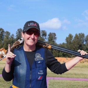 An athlete holds her shotgun while holding up the number one with her finger.